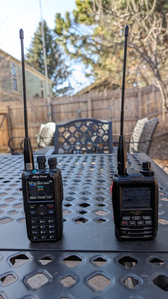 Anytone 878 and Yaesu FT-5DR handheld radios sitting on a patio table, both set to receive APRS data